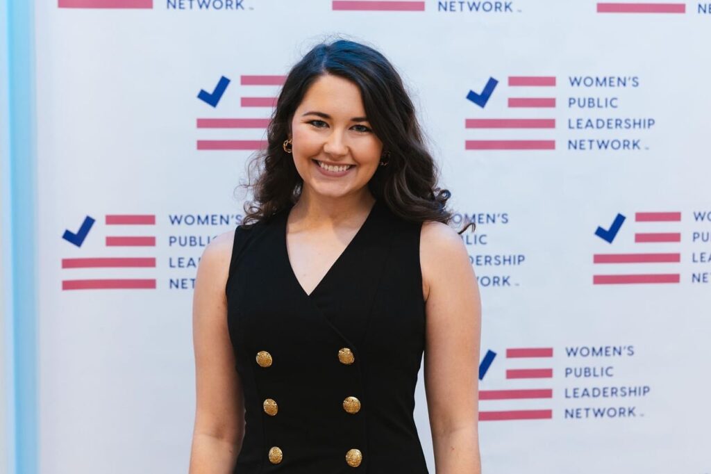 A woman in a black sleeveless outfit with brass buttons stands in front of a background that says Women's Public Leadership Network.