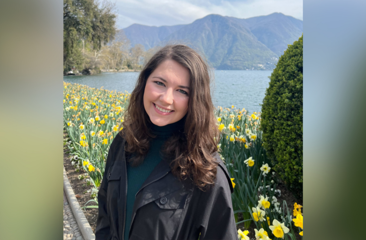 A young woman wearing a green shirt and black jacket poses for a photo, with daffodils, a lake and a mountain in the background.