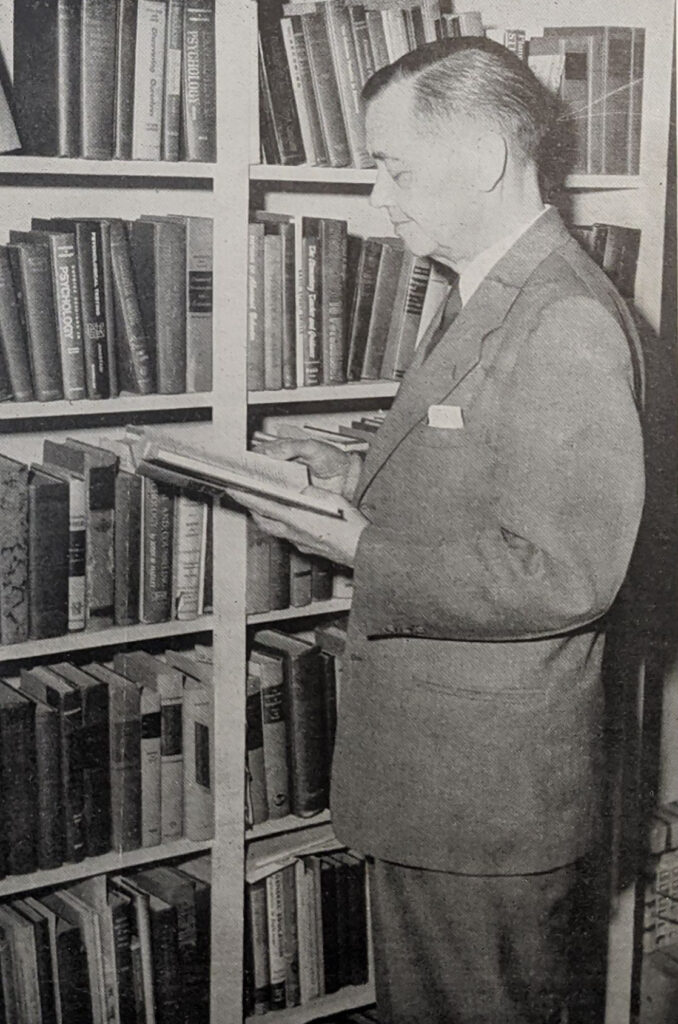 black and white photo of a man wearing a suit and standing next to a book shelf while looking at an open book in his hands