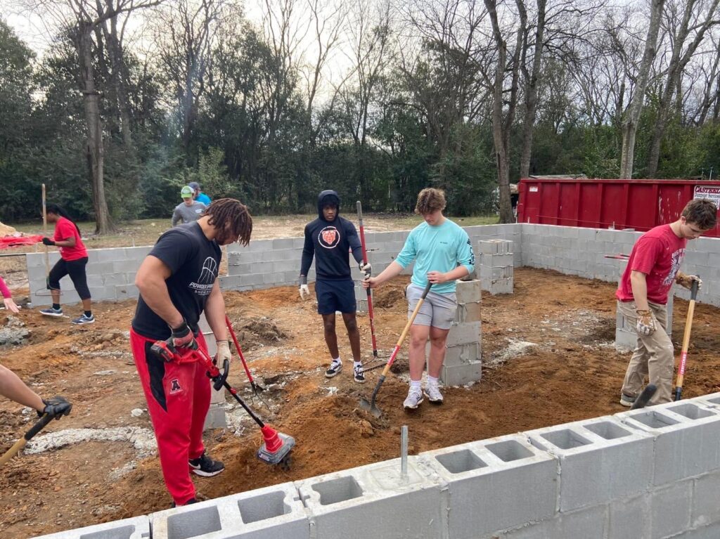 Several male college students hold shovels and dig in the dirt, with a border of concrete blocks outlining a home around them.