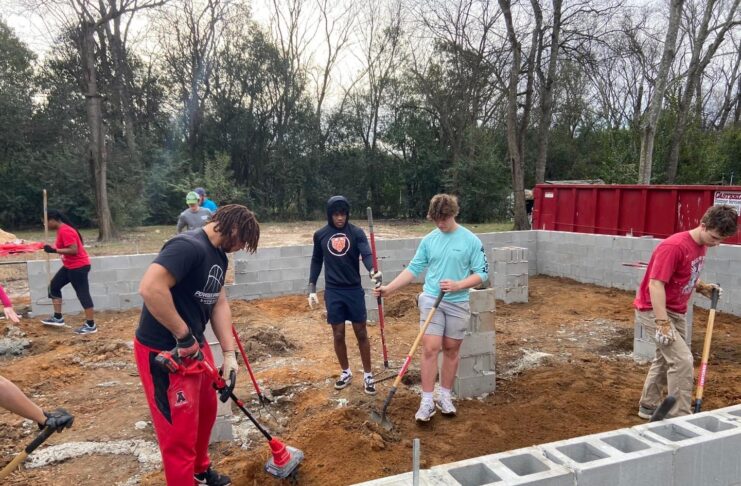 Several male college students hold shovels and dig in the dirt, with a border of concrete blocks outlining a home around them.