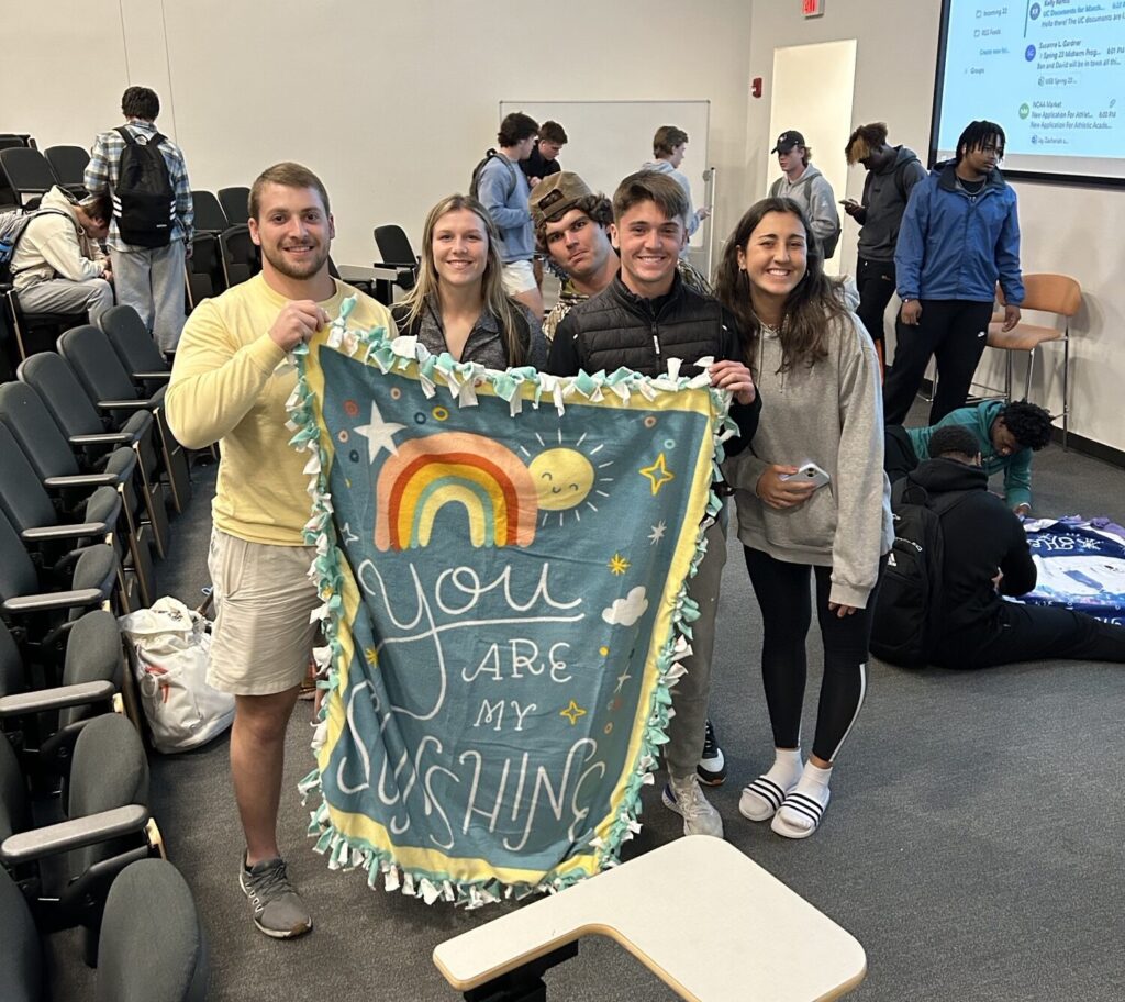 Several students in a lecture room hold up a blanket they made with a rainbow that says "You are my sunshine."
