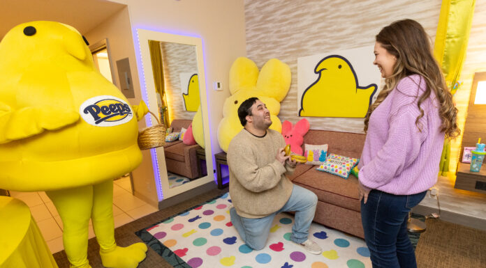 Mercer librarian’s Peeps-themed engagement gains national attention A man gets on one knee to propose while a woman stands in front of him. A yellow chick mascot stands beside him, holding an Easter basket. They stand in a room decorated with bright chick and bunny designs.