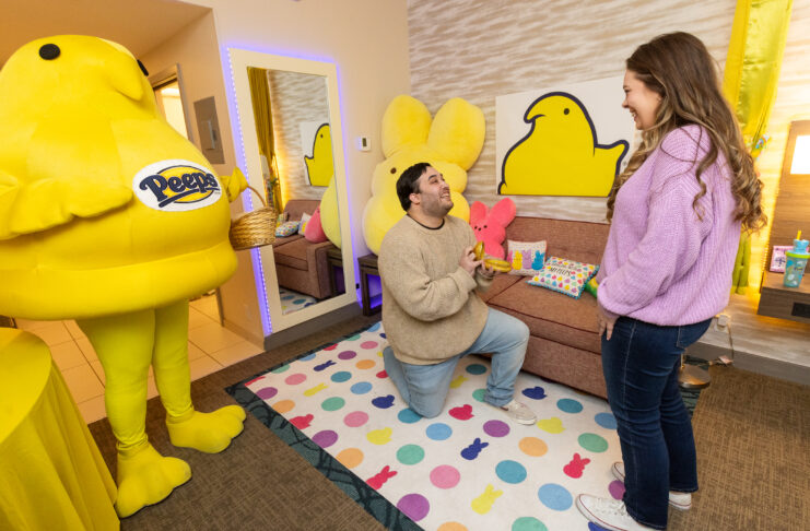 A man gets on one knee to propose while a woman stands in front of him. A yellow chick mascot stands beside him, holding an Easter basket. They stand in a room decorated with bright chick and bunny designs.