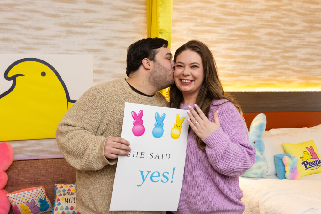 A man holds a sign that says "she said yes" while kissing a woman on the cheek who is holding up her hand to show an engagement ring.