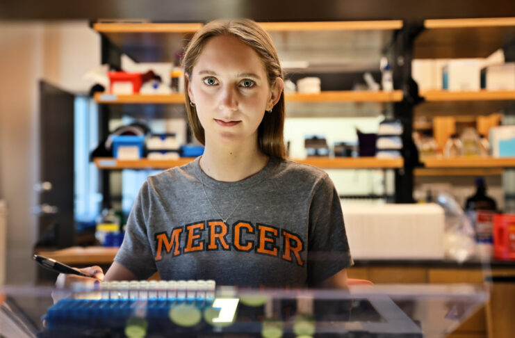 a female student wearing a mercer shoot looks at the camera. a lab is in the background
