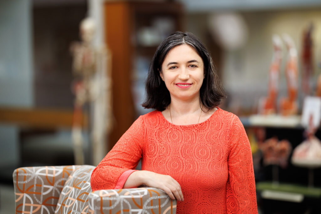 Dr. Ilana Chefetz Menaker is smiling while wearing an orange shirt. Dr. Menaker is standing next to a modern chair, with anatomical models in the background, suggesting an educational or medical setting.