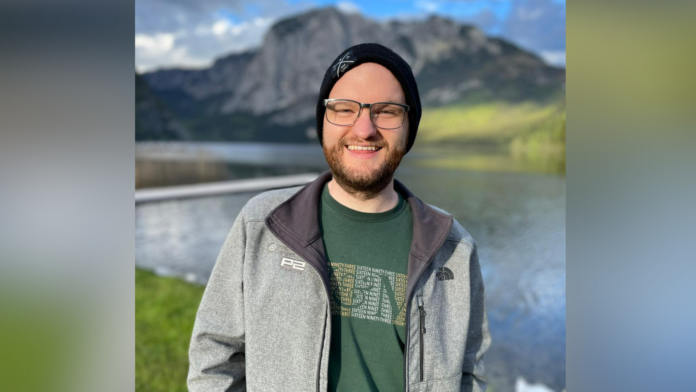 A man in a knit hat, green shirt and gray zip-up jacket is shown in front of a lake and mountain in the Austrian Alps.