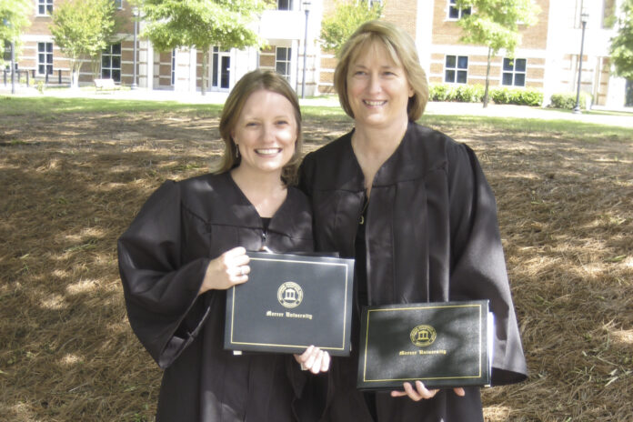 mom and daughter smile at the camera while wearing black graduation robes and holding their diplomas
