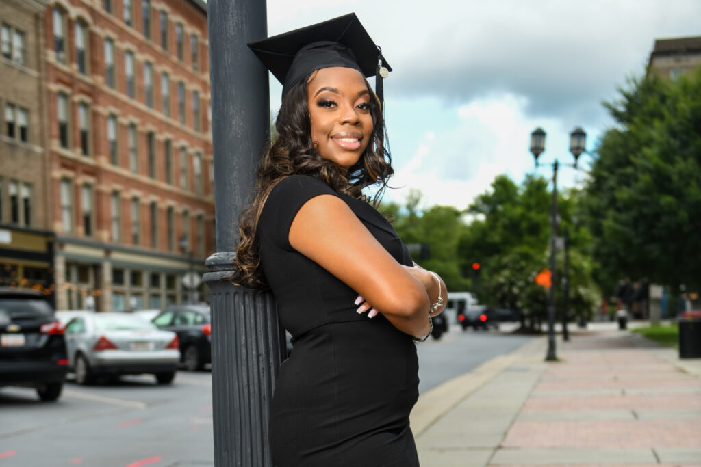 woman wearing a black dress and graduation cap leans against a pole in downtown Macon