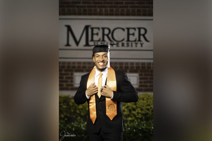 man wears graduation regalia while standing in front of a mercer university sign