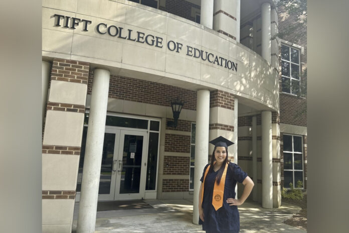 a woman wearing a black dress, orange stole and graduation cap stands in front of the Tift College of Education building