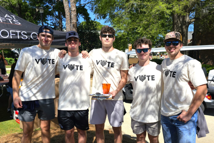 Five members of Mercer's men's lacrosse stand outside wearing matching white shirts that say "Vote" on the front. The "O" in vote is the Mercer bear head. The individual in the middle is holding a clipboard. On top of the clipboard is a cup with pens in it.