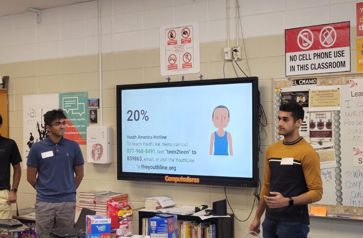 Two male students stand on either side of a wall-mounted TV with a presentation displayed.