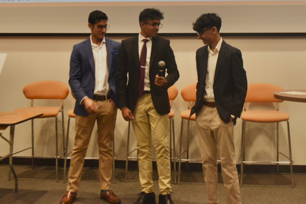 Three male students in khakis and nice jackets hold a microphone for a presentation
