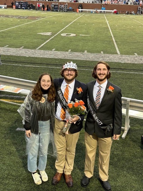 A female college student in jeans, a sweater and a rain poncho stands next to her two brothers, who are in Homecoming court and one wearing a crown after being named king, wearing suits and black sashes.