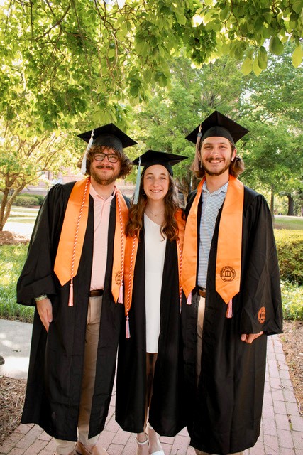 Two brothers and a sister post for a photo with their graduation caps and gown and orange stoles.