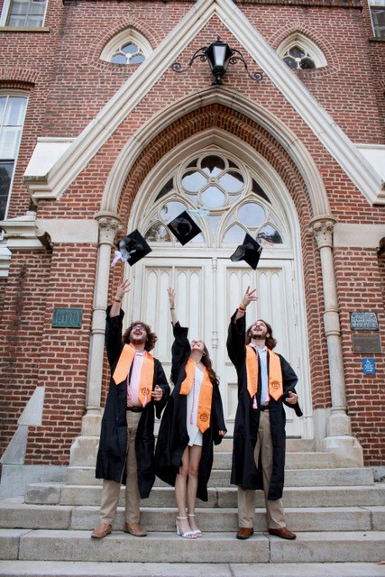 Two brothers and a sister wearing graduation robes and orange stoles throw their caps in the air, on the steps of a brick building with white entry doors. 