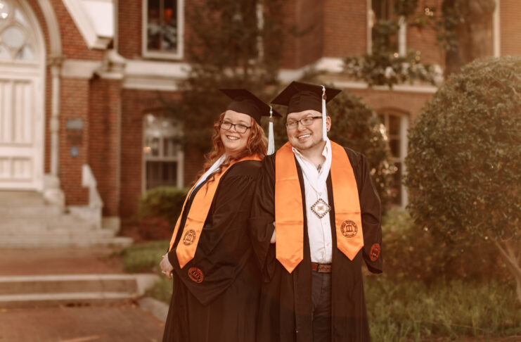 man and woman wearing graduation regalia stand in front of admin building