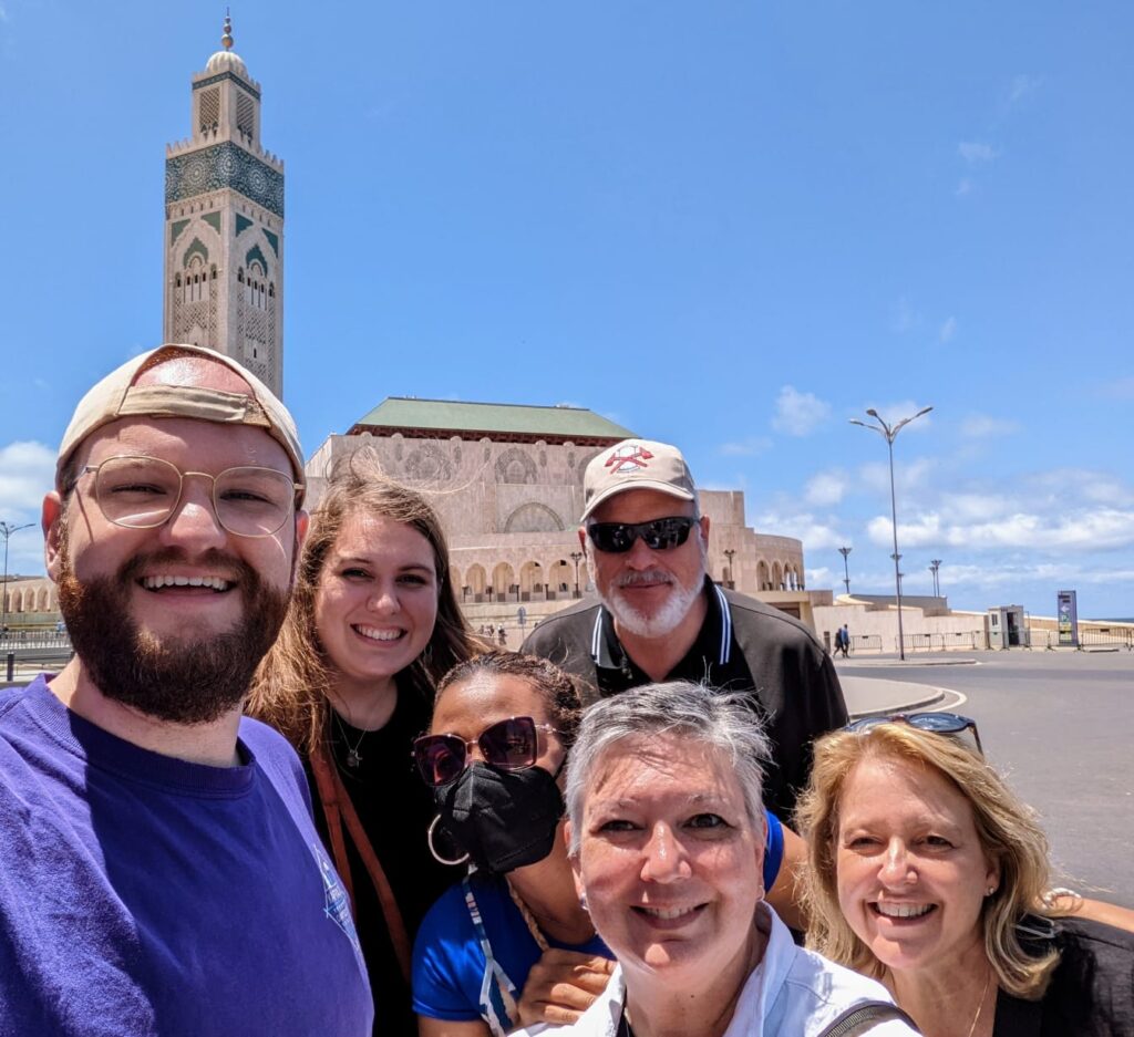 A group of six adults stand take a selfie in front of large, historic buildings in Morocco.