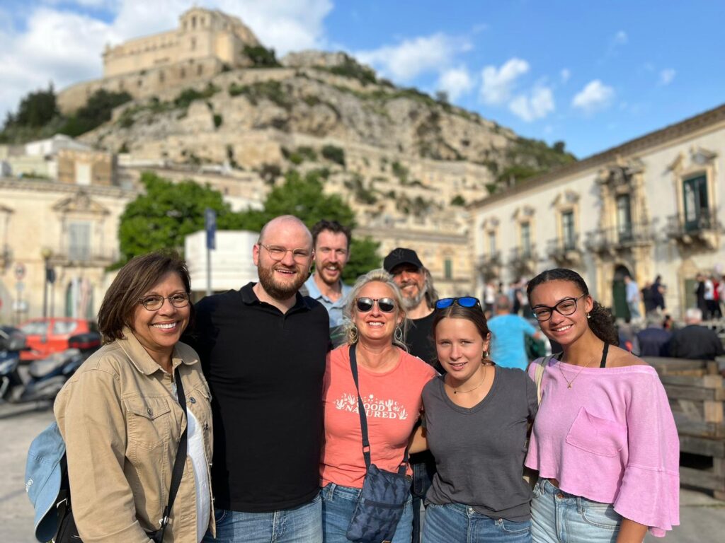 A group of seven adults take a photo as a historic buildings loom on a mountain behind them in Sicily.