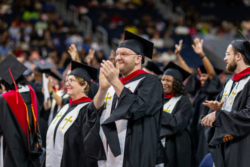 A photo showing a group of theology graduates in black robes and caps and white stoles with gold crosses are shown during commencement. The man in the middle claps and smiles.