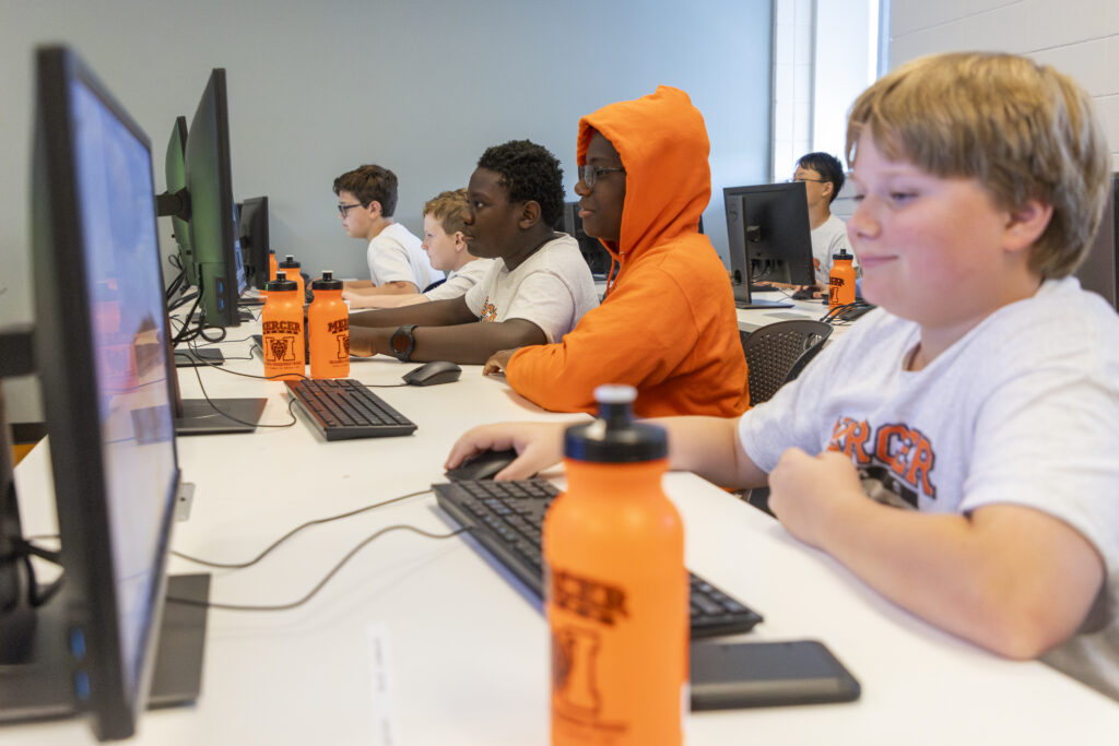 A group of students at a computer lab desks engaging intently with their screens during a class session, with some wearing Mercer-branded shirts alongside orange water bottles featuring the same logo.