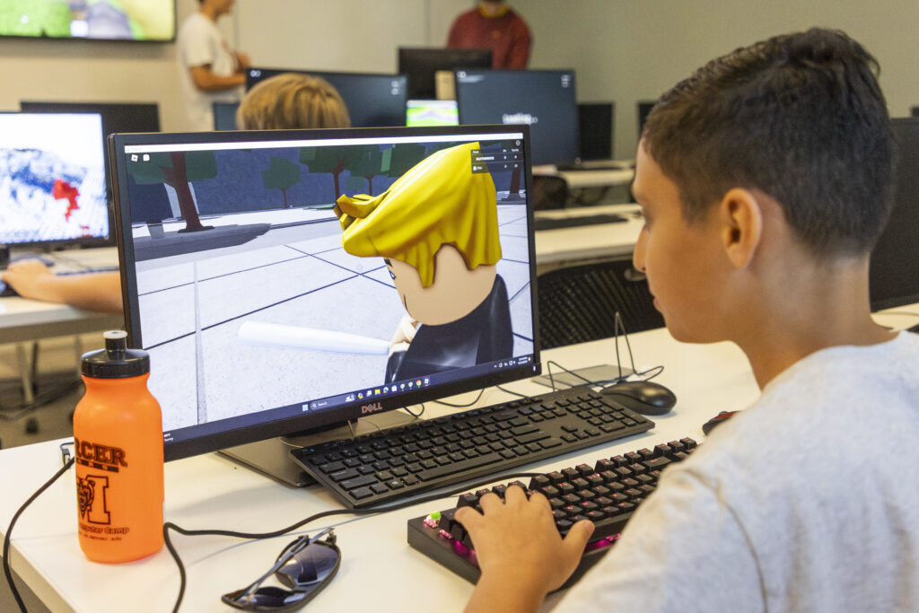 A person using a computer displaying a Roblox game scene in a classroom setting. An orange water bottle and sunglasses are placed next to the keyboard.