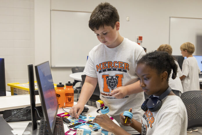 Two children wearing shirts with the Mercer logo are engaged in a robotics workshop inside a classroom environment. They assemble components of a robot while looking at instructions on a computer screen.