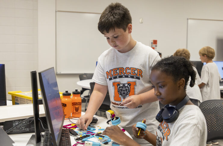 Two children wearing shirts with the Mercer logo are engaged in a robotics workshop inside a classroom environment. They assemble components of a robot while looking at instructions on a computer screen.