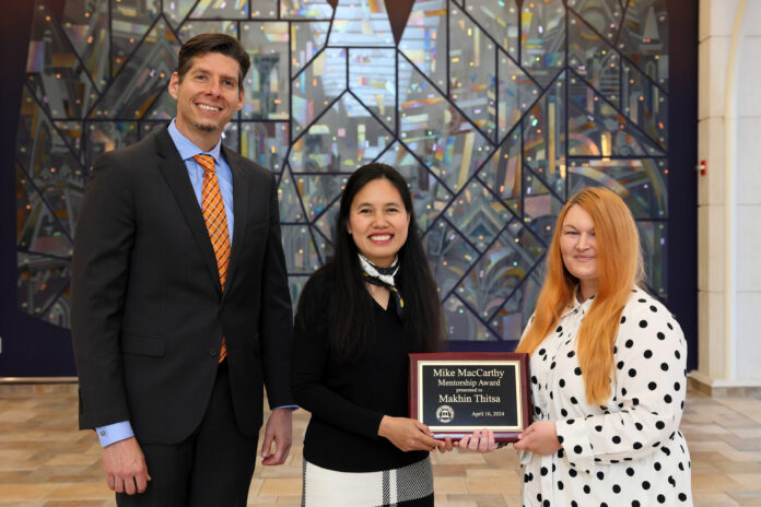 Three people standing in front of a colorful stained-glass background. The woman in the middle is a holding a plaque that reads 'Mike MacCarthy Mentorship Award presented to Makhin Thitsa, April 16, 2024.'
