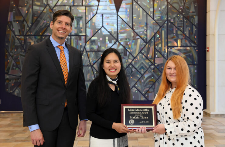 Three people standing in front of a colorful stained-glass background. The woman in the middle is a holding a plaque that reads 'Mike MacCarthy Mentorship Award presented to Makhin Thitsa, April 16, 2024.'