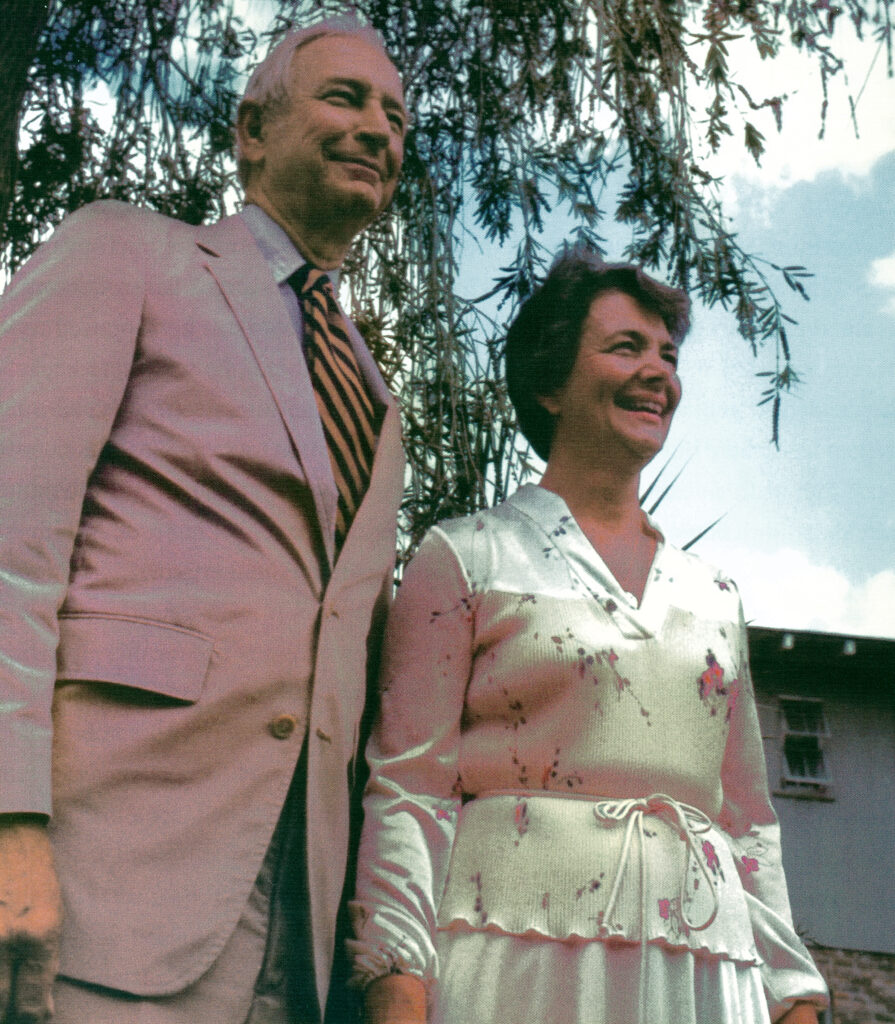 A man in a suit and tie and a woman in a flowery silk dress standing under a willow tree, smiling and looking towards the distance.