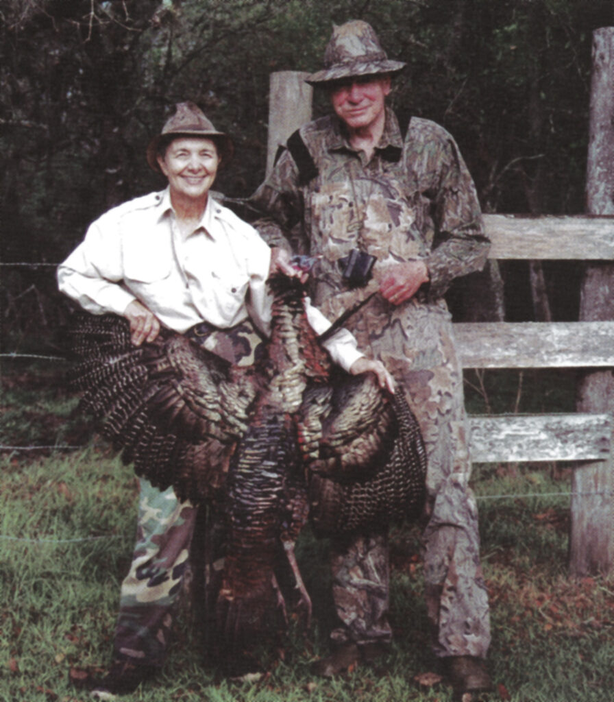 A woman and man wearing hats and outdoor clothing hold a large turkey. They stand next to a wooden fence in a grassy area.