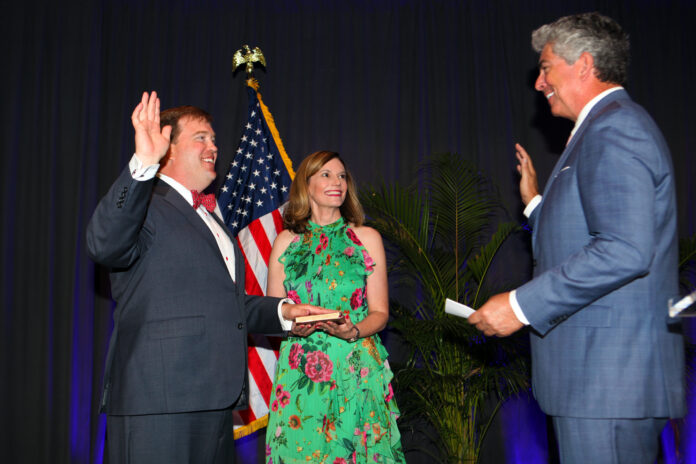 A man wearing a navy blue suit and red bow tie is raising his right hand, taking an oath. A woman in a green floral dress is standing beside him, holding a book on which the man places his left hand. Another man, dressed in a blue suit, is administering the oath, also with his right hand raised. An American flag and some green plants are visible in the background.