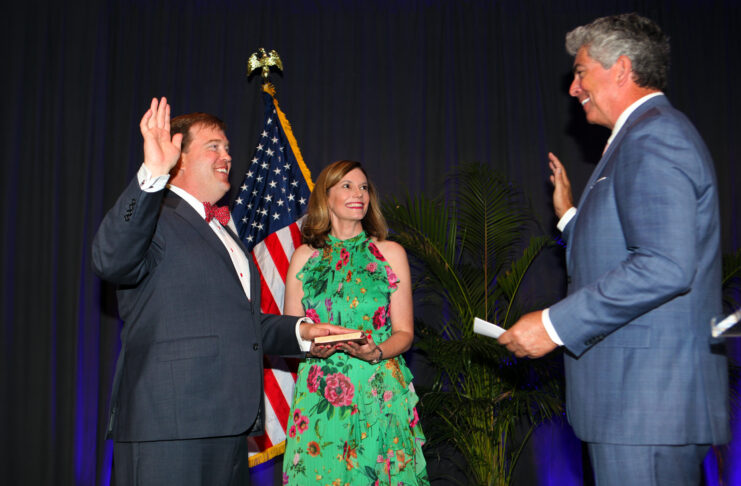 A man wearing a navy blue suit and red bow tie is raising his right hand, taking an oath. A woman in a green floral dress is standing beside him, holding a book on which the man places his left hand. Another man, dressed in a blue suit, is administering the oath, also with his right hand raised. An American flag and some green plants are visible in the background.