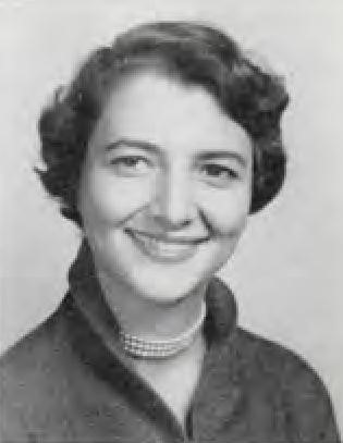 Black and white portrait of a smiling woman with short curly hair, wearing a collared shirt and a pearl necklace.