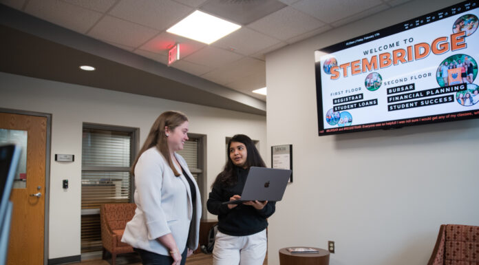 Mercer staff are leaders in Georgia financial aid association Two individuals are engaging in a discussion in a modern office lobby, with one holding a laptop; in the background, a digital display shows information about services such as financial planning and student loans.