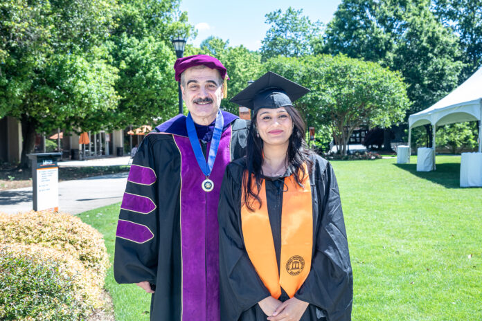 A female graduate in cap and gown, wearing an orange stole, smiles next to a man in an academic ceremonial robe and hat.  They are outdoors on a sunny day, with green trees and grass in the background.