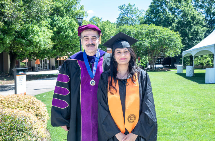 A female graduate in cap and gown, wearing an orange stole, stands smiling next to a man in a ceremonial academic robe and hat. They are outdoors on a sunny day with green trees and grass in the background.