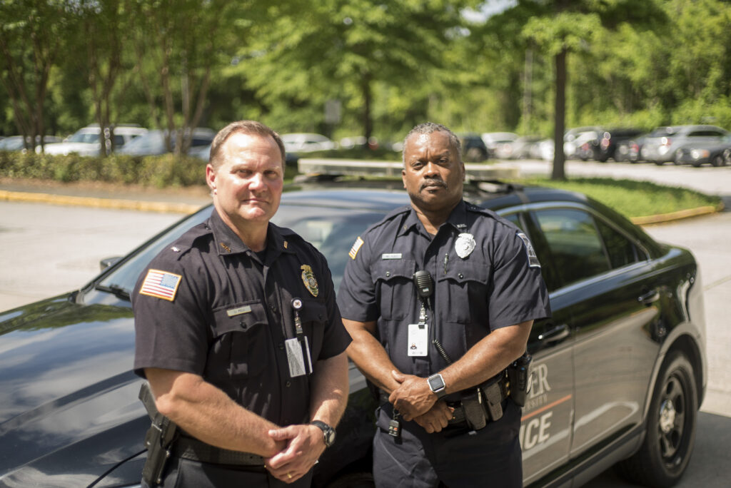 Two police officers standing in front of a patrol car in a parking lot with trees in the background. Both are wearing badges and the car has "Mercer Police" written on it. They appear focused and professional.