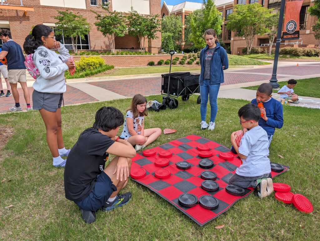Four children sit around a large game of checkers in the grass outside, while two college students stand up behind them, with brick academic buildings in the background.