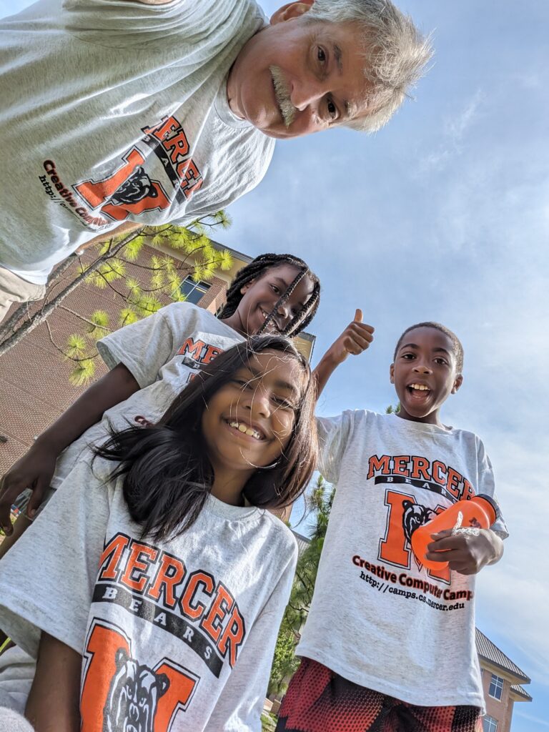 A professor and three children, all in matching white T-shirts with orange letters that say "Mercer Bears Creative Computer Camps" and have the Mercer logo.
