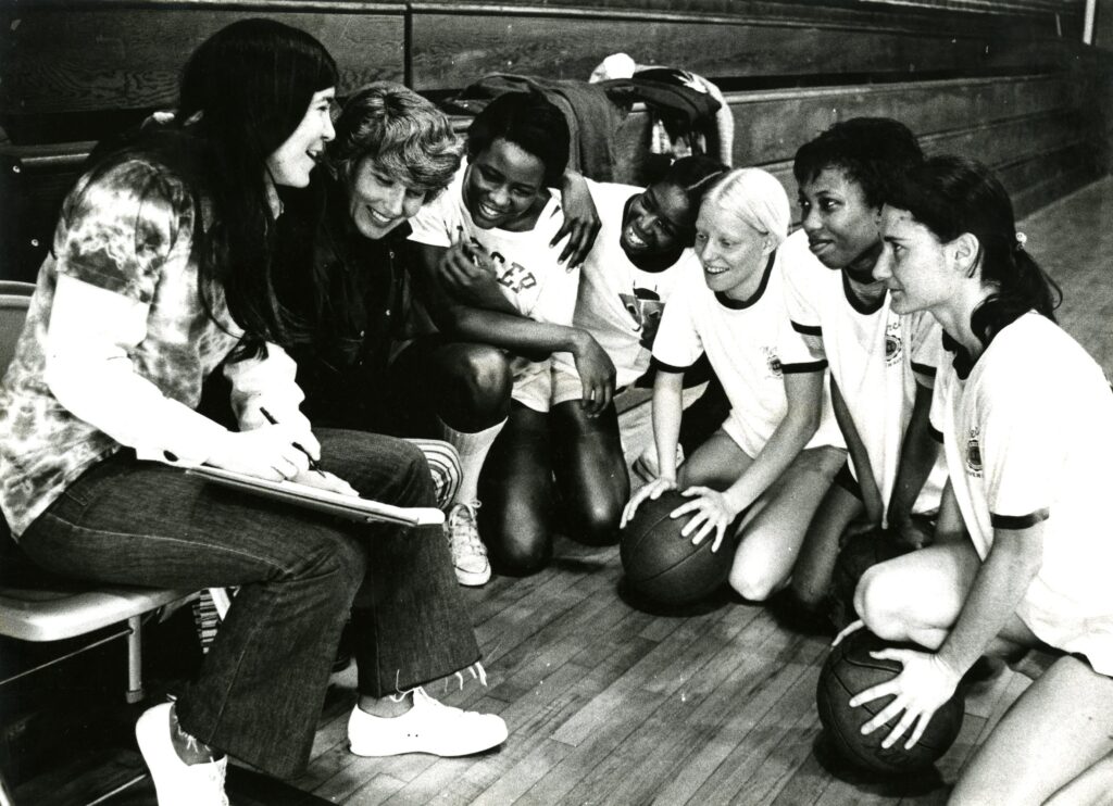 Group of women's college basketball players kneel on the gym floor, smiling and interacting joyfully with their coach, who is holding a clipboard.