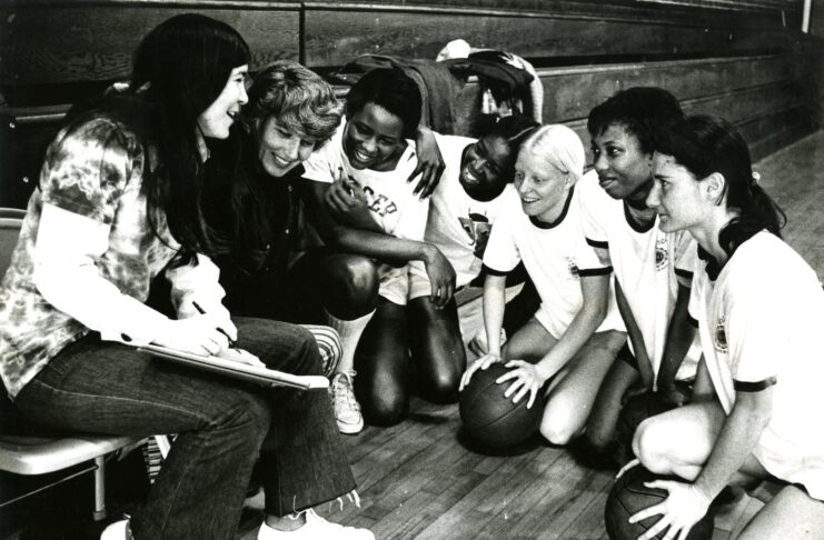 Group of women's college basketball players kneel on the gym floor, smiling and interacting joyfully with their coach, who is holding a clipboard.