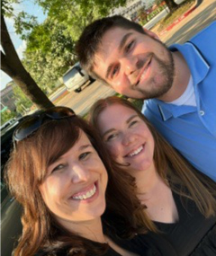 A selfie of three people outdoors in a parking lot. The person on the left is a woman with shoulder-length brown hair, wearing sunglasses on her head and a black top, smiling at the camera. In the middle is another woman with long reddish-brown hair, also smiling, wearing a black top. On the right is a man with short dark hair and a beard, smiling, and wearing a light blue polo shirt. The background includes trees, parked cars, and a clear sky.