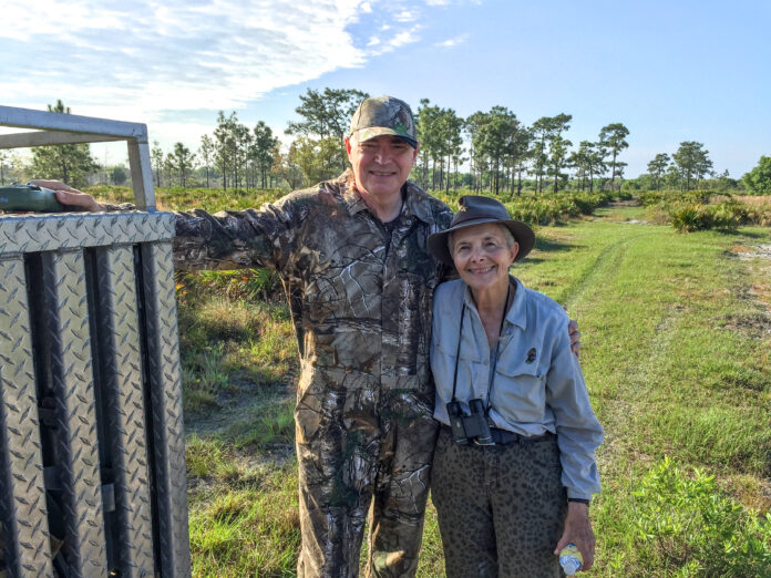 A man and woman in hunting clothing and hats are shown in front of a grassy and tree-filled area.