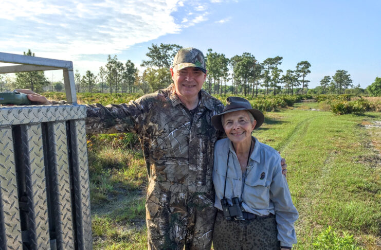 A man and woman in hunting clothing and hats are shown in front of a grassy and tree-filled area.