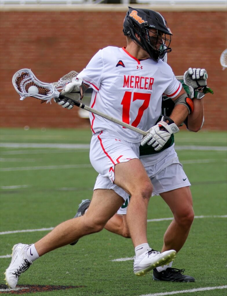 A man in a white jersey with the word "Mercer" and the number 17 in orange holds a lacrosse stick while running down the field.