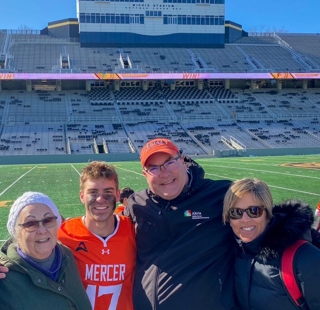 A male student in an orange jersey is pictured with his grandmother, father and mother with a stadium in the background.